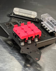 A close-up of a black metal fuse holder or terminal assembly with three bright pink fuse bodies in the center and clear plastic fuse housings on a brushed metal surface.