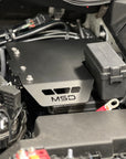 A close-up of an MSD brand black metal engine cover installed in a vehicle's engine bay, alongside a gray fuse box.
