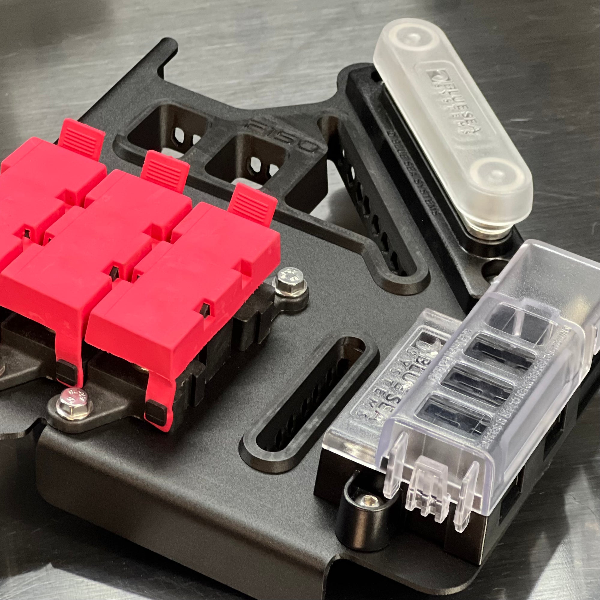 Close-up of a black metal mounting bracket with bright pink connector blocks, a translucent white cylindrical component, and a clear plastic multi-pin socket on a metallic gray surface.