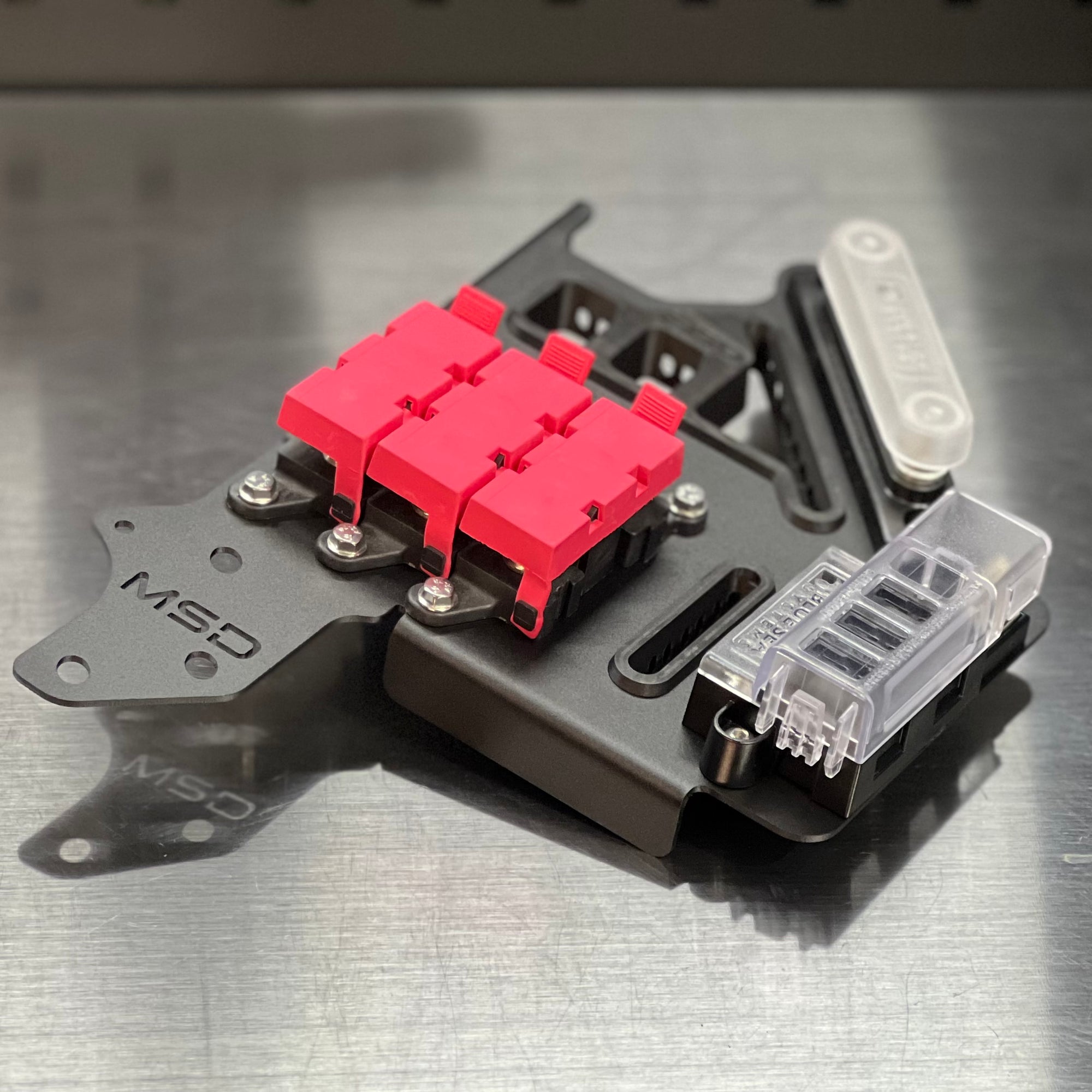 A close-up of a mechanical component or hardware part with a bright pink plastic section, black metal base, silver screws, and a clear plastic cover, photographed on a brushed metal surface.