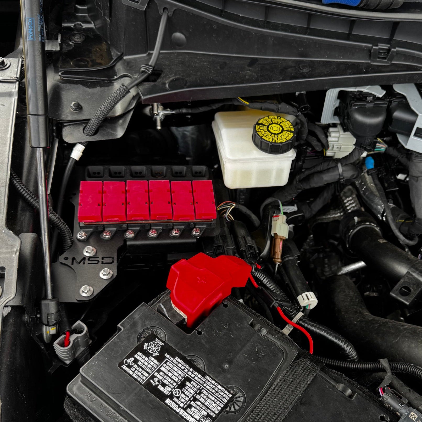 Close-up of a car engine bay showing the black automotive battery, red terminal cover, white reservoir tank, and surrounding engine components.