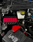 Close-up of a car engine bay showing the black automotive battery, red terminal cover, white reservoir tank, and surrounding engine components.