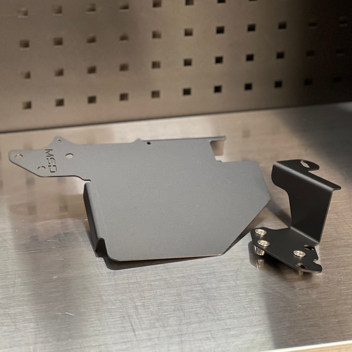A dark gray metal engine cover shield and a black mounting bracket with screws, displayed on a brushed metal surface against a perforated metal background.