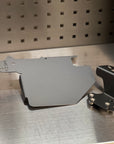 A dark gray metal engine cover shield and a black mounting bracket with screws, displayed on a brushed metal surface against a perforated metal background.