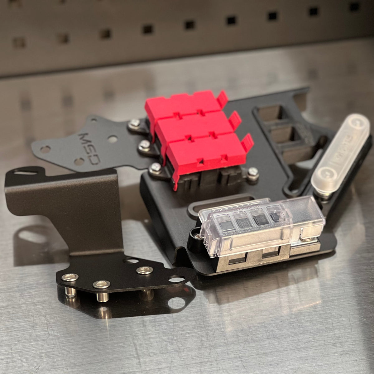 A close-up of a custom metal electronic component assembly, featuring a black anodized chassis with a bright red plastic connector block, a clear plastic fuse holder, and mounting brackets, resting on a brushed metal surface.