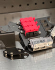 A close-up of a custom metal electronic component assembly, featuring a black anodized chassis with a bright red plastic connector block, a clear plastic fuse holder, and mounting brackets, resting on a brushed metal surface.