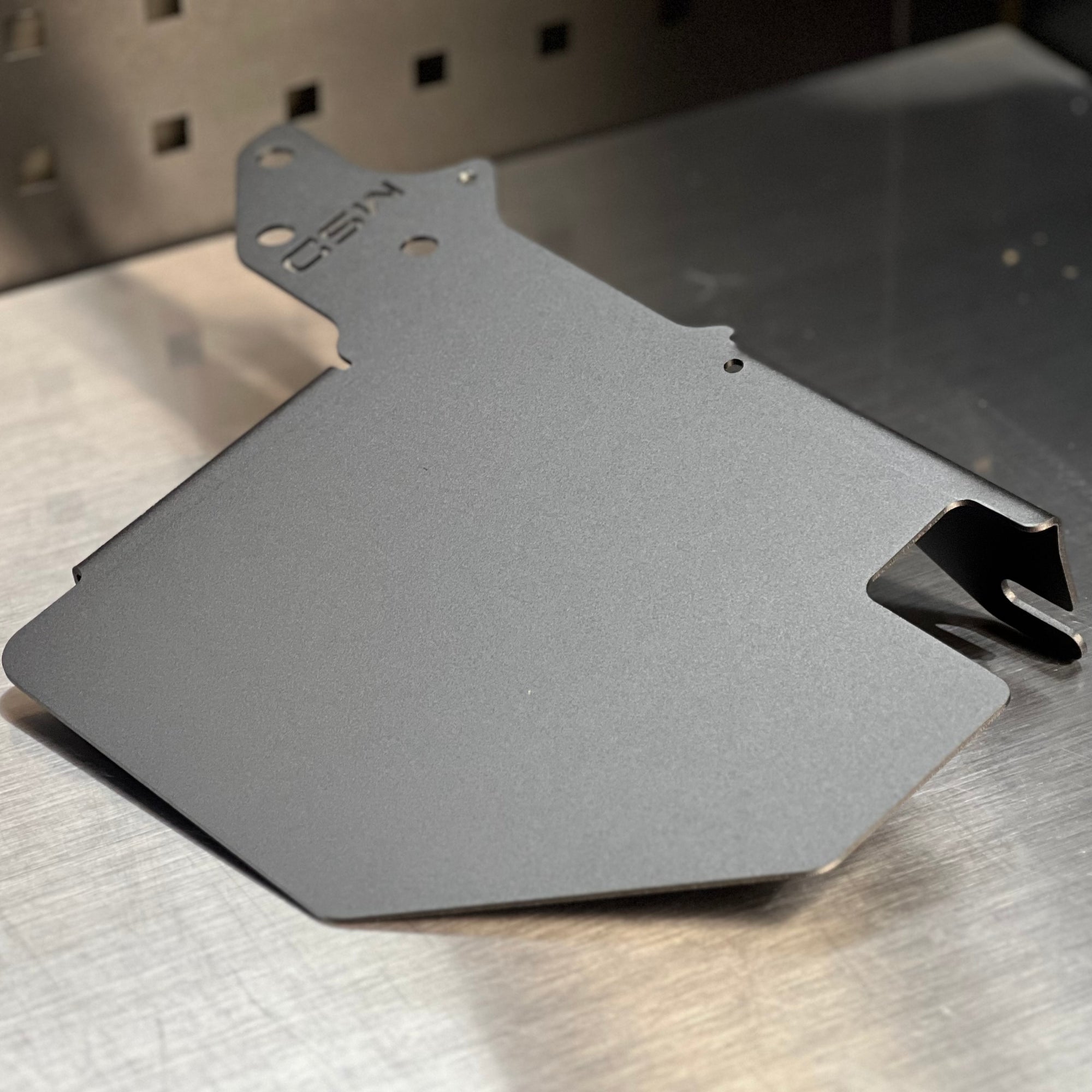 A matte dark gray metal mounting plate or bracket with pre-drilled holes and a stamped 'CSN' logo, resting on a brushed metal workbench.