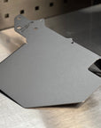 A matte dark gray metal mounting plate or bracket with pre-drilled holes and a stamped 'CSN' logo, resting on a brushed metal workbench.