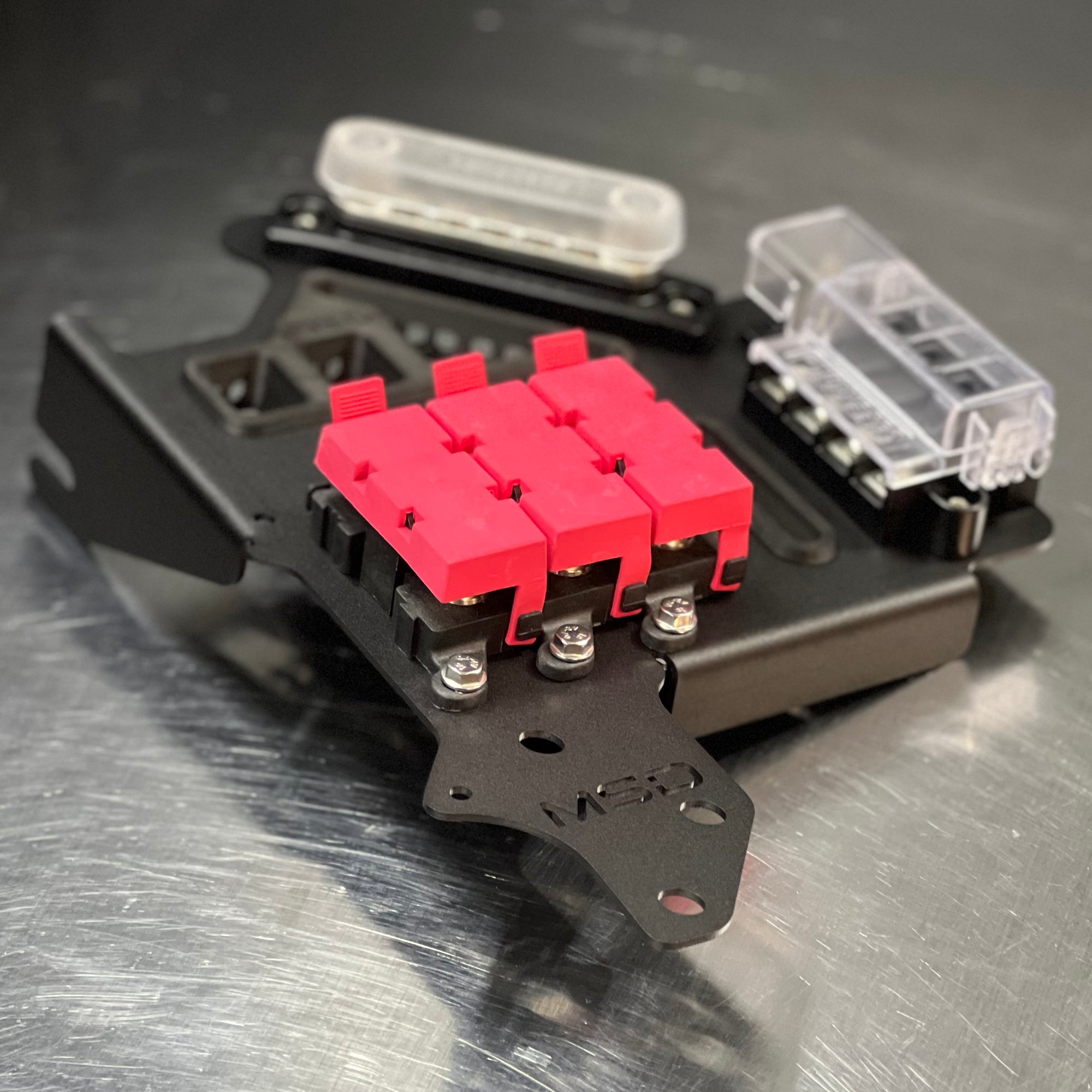 A close-up of a black metal fuse holder or terminal assembly with three bright pink fuse bodies in the center and clear plastic fuse housings on a brushed metal surface.