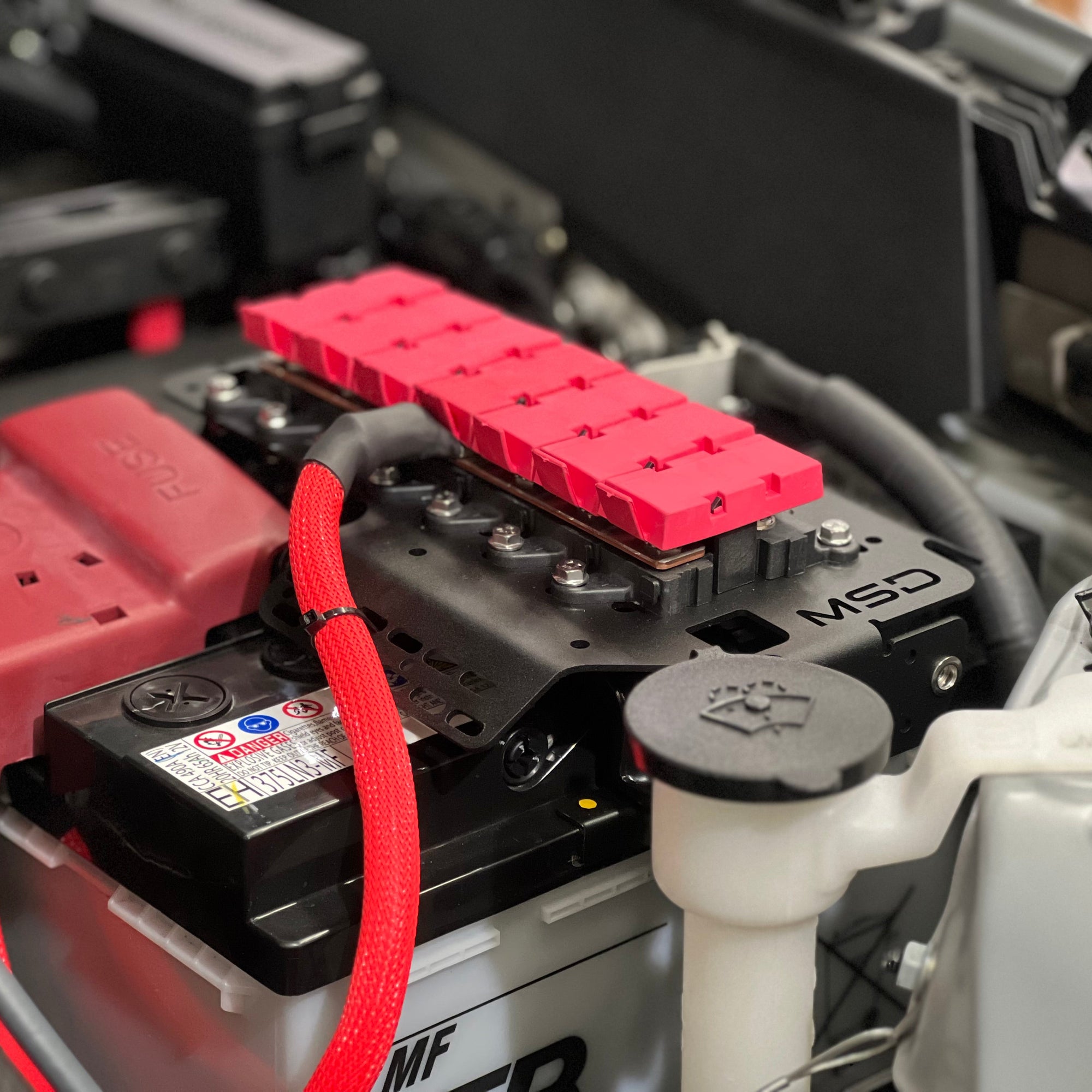 A close-up of a car battery with a bright red protective cover and matching red cable sleeve, installed inside a vehicle engine bay alongside other mechanical components.