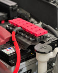 A close-up of a car battery with a bright red protective cover and matching red cable sleeve, installed inside a vehicle engine bay alongside other mechanical components.