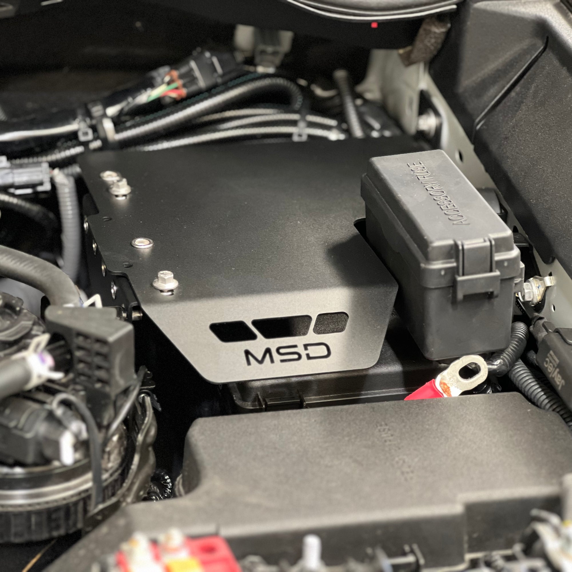 A close-up of an MSD brand black metal engine cover installed in a vehicle's engine bay, alongside a gray fuse box.