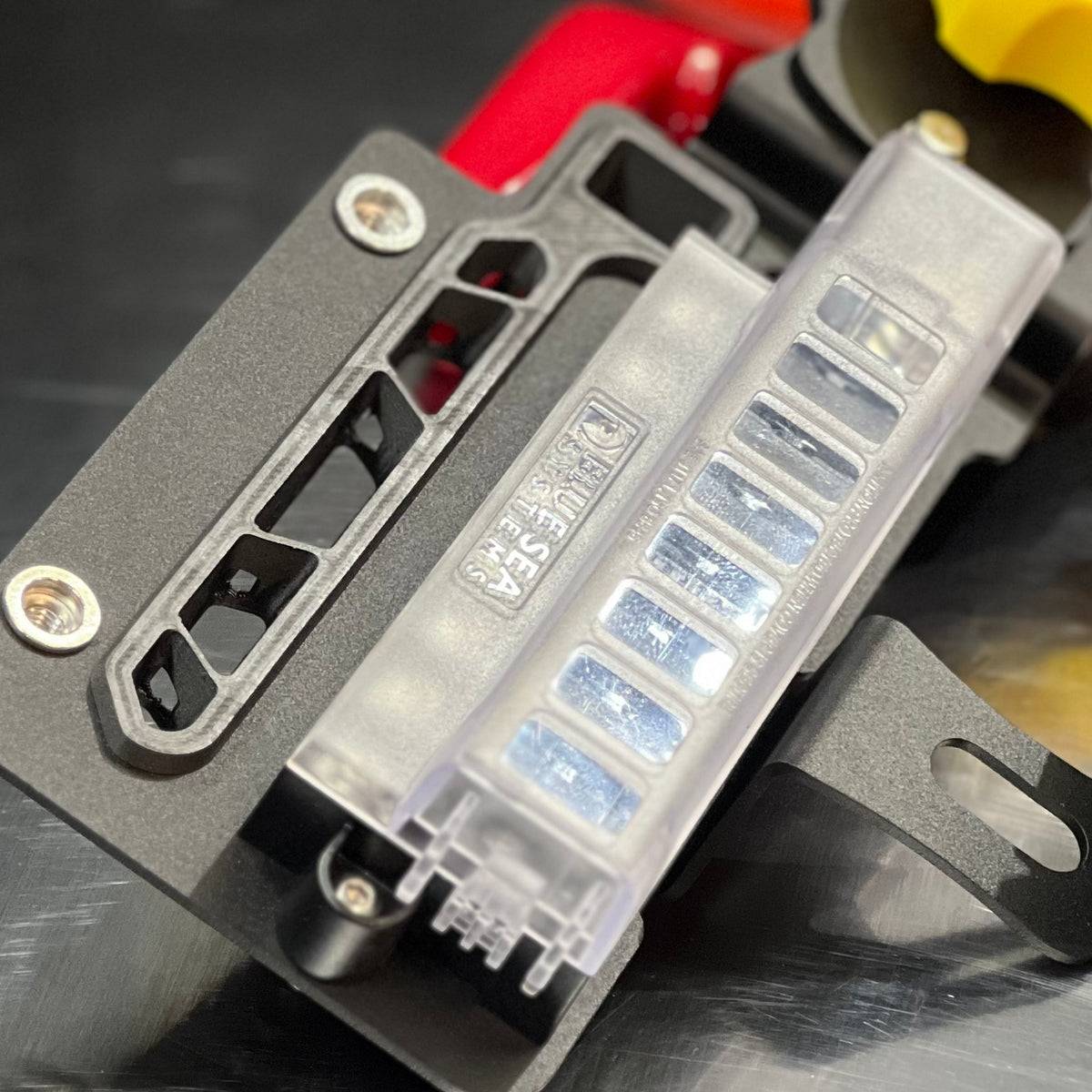 Close-up of a black metal mounting bracket with a clear plastic fuse holder containing multiple blue-tinted fuses, photographed on a dark reflective surface.