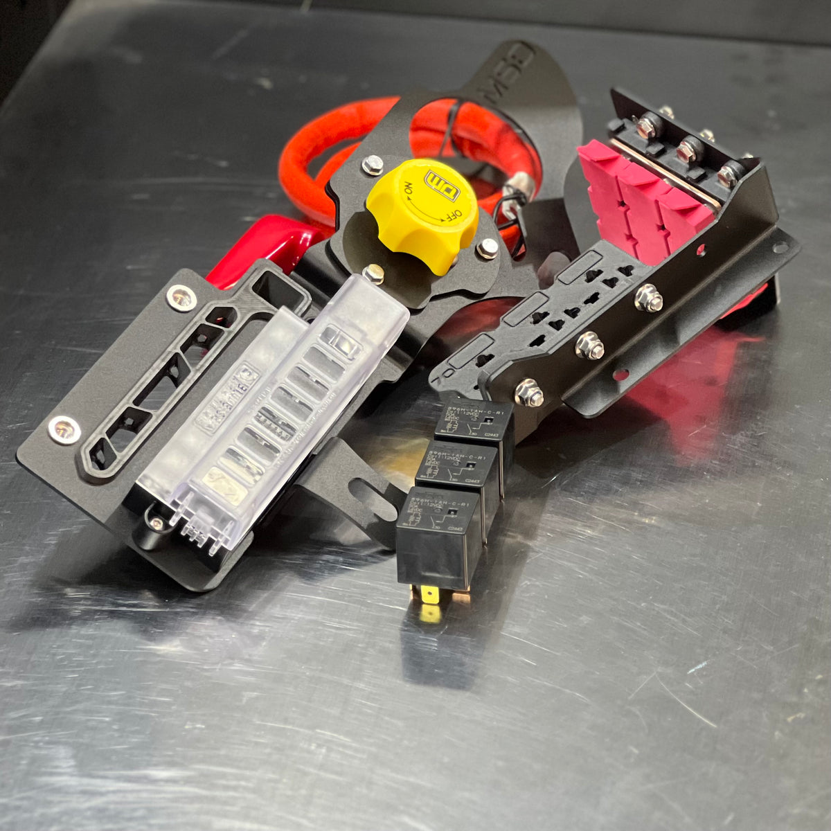 A close-up of a custom-built metal electrical component or custom motor assembly, featuring a black anodized chassis, red and orange insulated wires, a yellow plastic knob, a clear plastic fuse holder, and black relay blocks on a scratched dark gray metal surface.