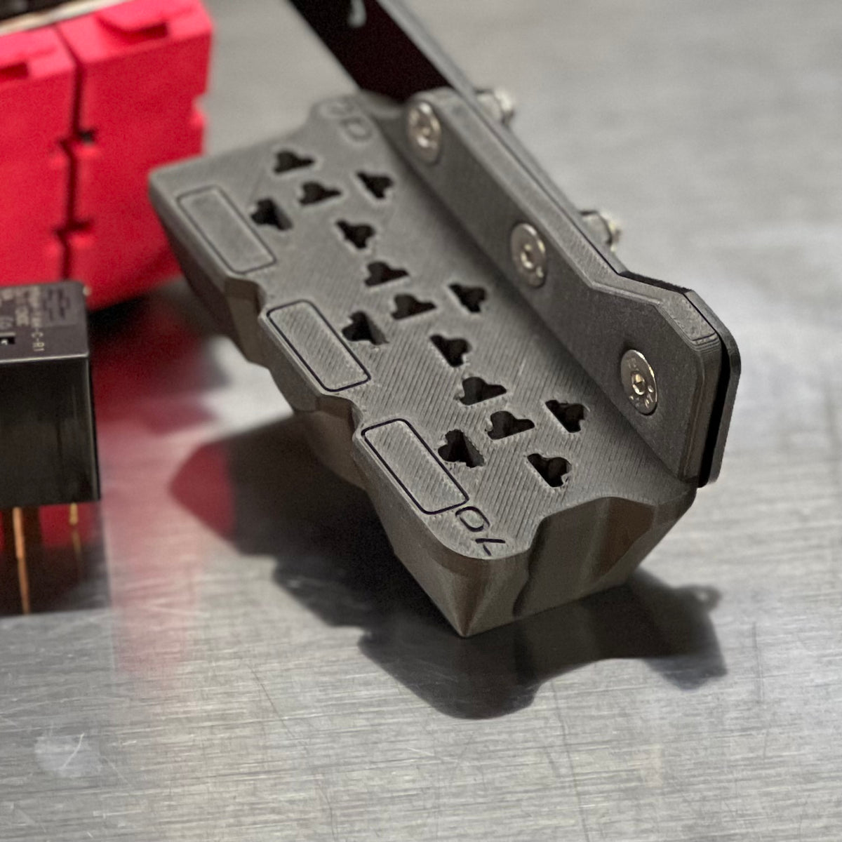 A close-up of a dark gray 3D-printed multi-plug power adapter or outlet cover, shown on a brushed metal surface with other electronic components partially visible in the background.