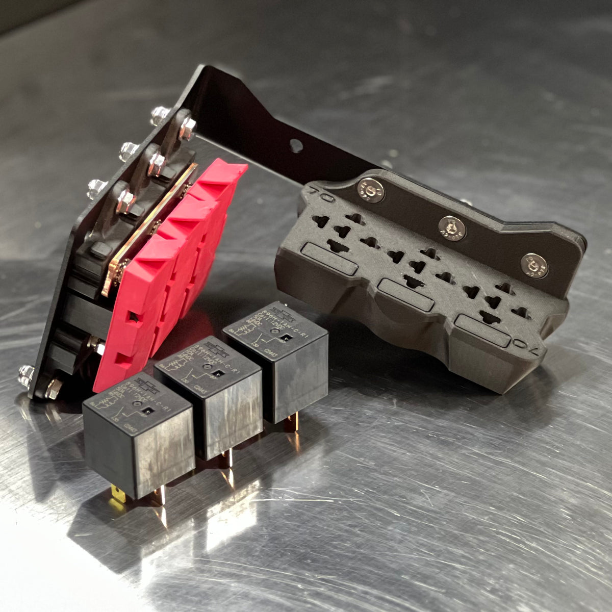 A set of industrial electrical components, including a black relay module with a prominent red terminal block, a black metal bracket, and three small black relays, displayed on a brushed metal surface.